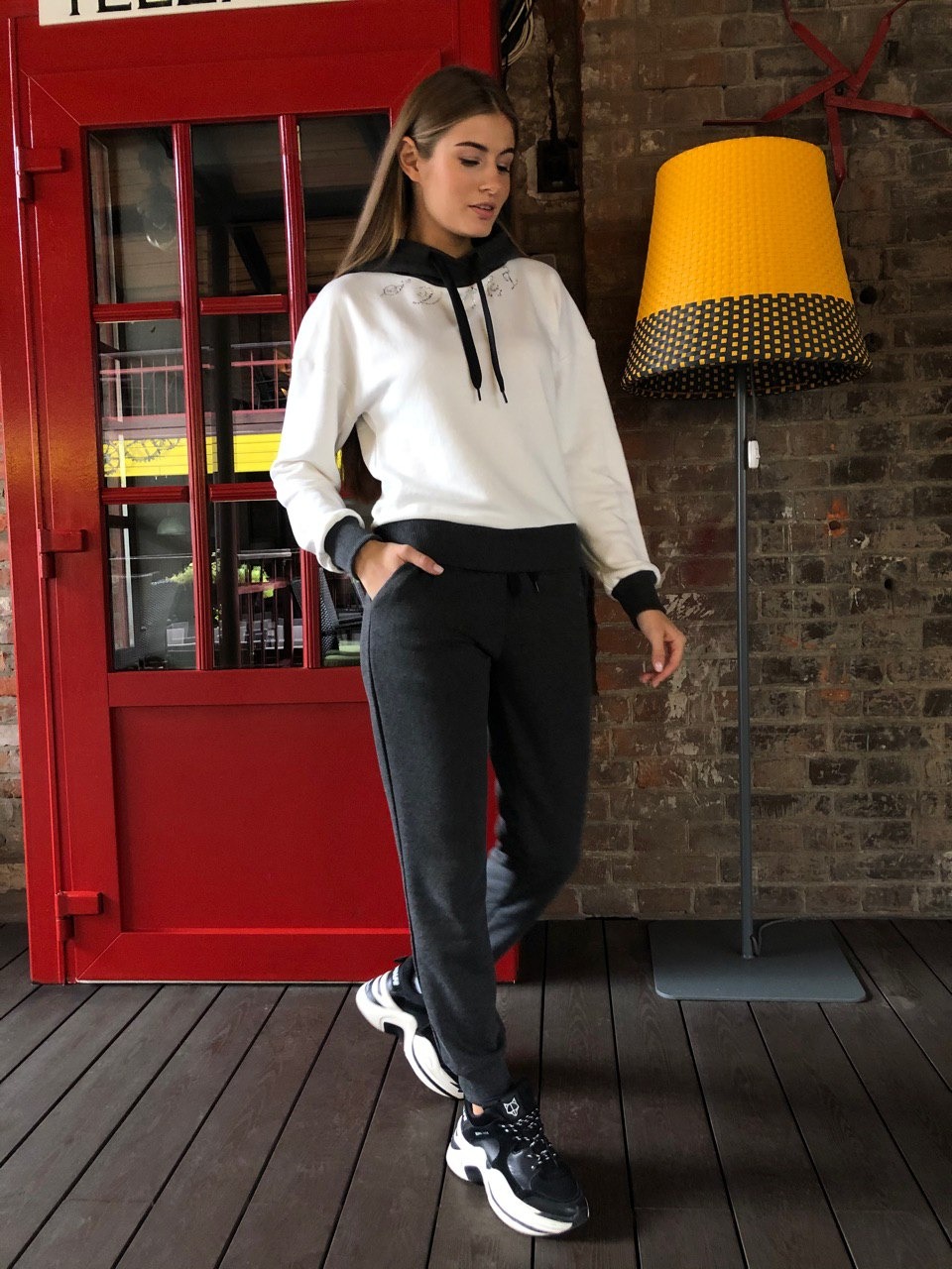 Kobieta prezentuje dresowy komplet: biała bluza z kapturem ozdobiona drobnymi aplikacjami i grafitowe spodnie dresowe, stojąc na drewnianej podłodze obok czerwonej budki telefonicznej i żółtej lampy.
