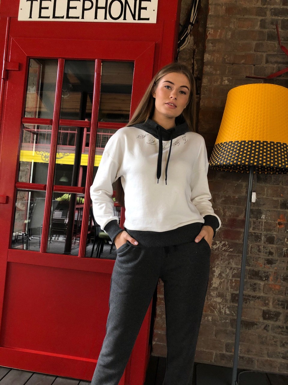 Młoda kobieta pozuje w dresie w studio z czerwoną budką telefoniczną i lampą z wikliny w tle.