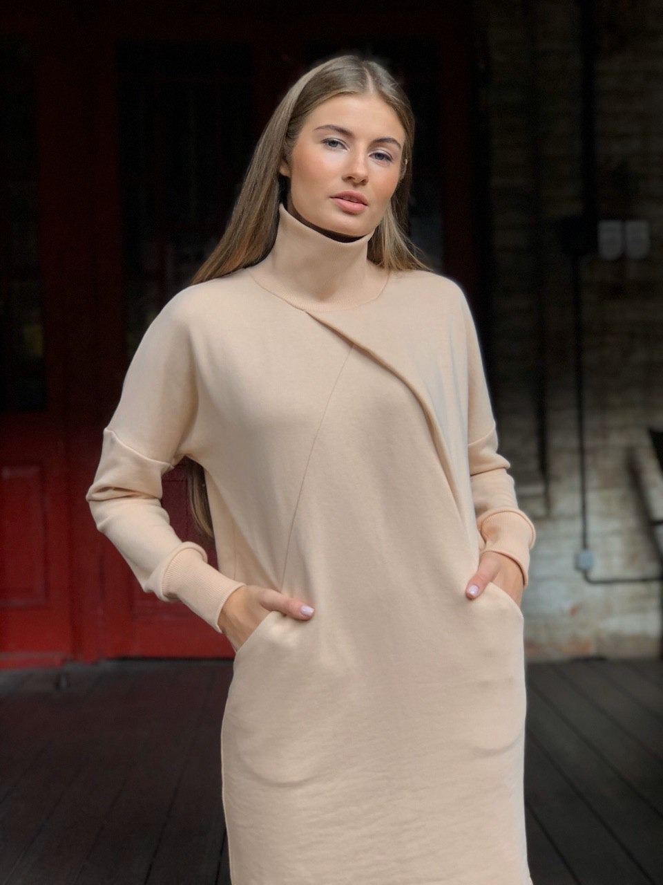 Portret kobiety w beżowej sukience z asymetrycznym dekoltem na tle czerwonych drzwi i ceglanej ściany.