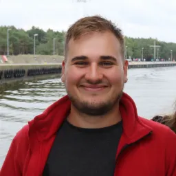 Uśmiechnięty mężczyzna w czerwonej polarowej kurtce na tle nabrzeża z wodą, drzewami i latarniami.