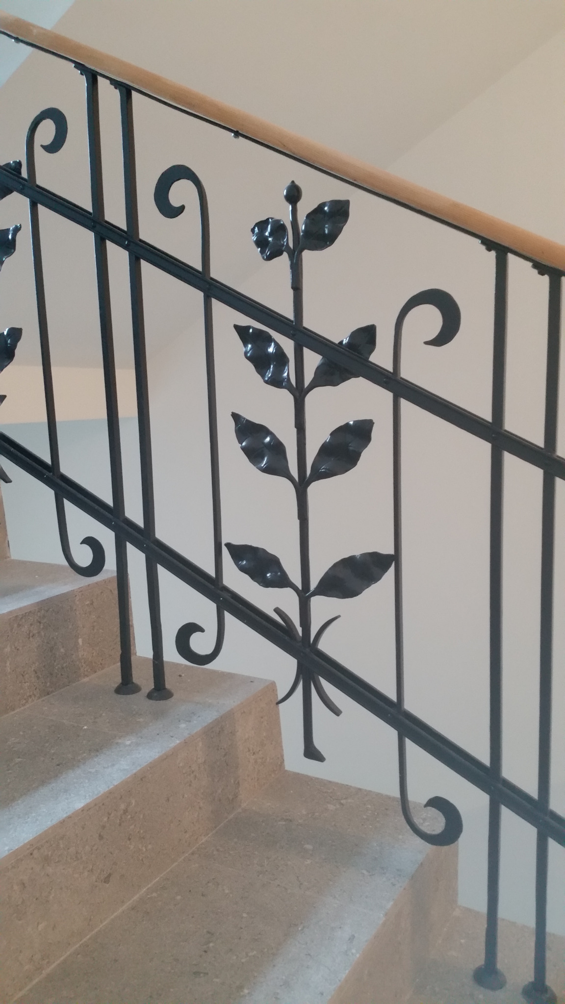 Detal metalowej balustrady schodowej z motywem roślinnym, czarne elementy kute na tle jasnych schodów i ściany.