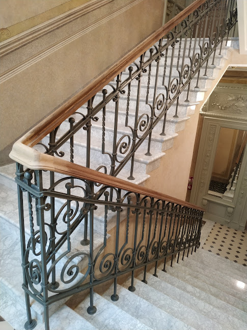 Zabytkowa, spiralnie zdobiona balustrada kuta na marmurowych schodach w budynku z elementami sztukaterii.
