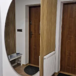 Aranżacja przedpokoju z owalnym lustrem, drewnianymi lamelami na ścianie, grzejnikiem i ceramiczną podłogą imitującą kamień.