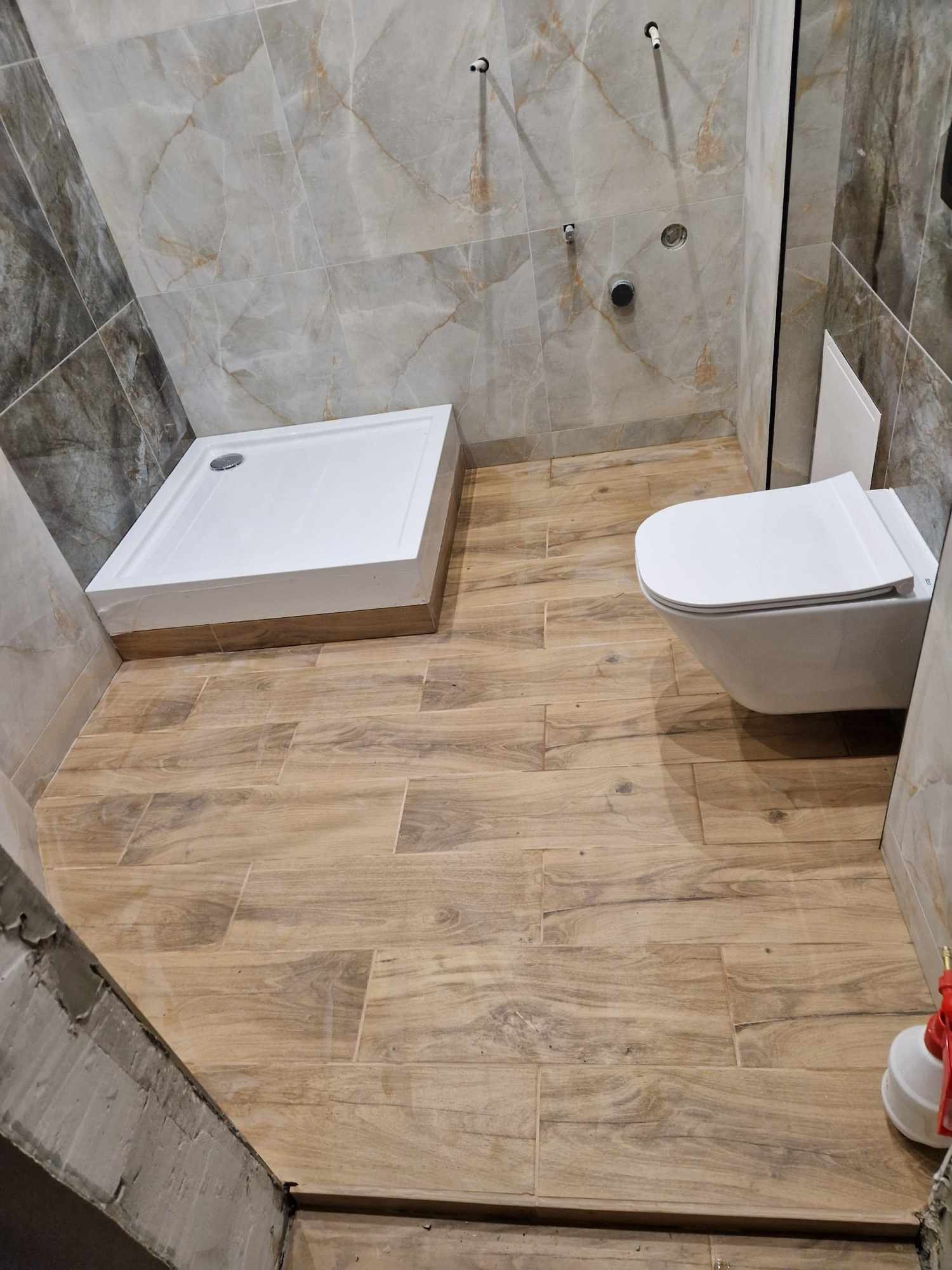 Nowoczesna łazienka z beżowymi płytkami imitującymi drewno na podłodze, biały brodzik i podwieszana toaleta, marmurowe płytki na ścianach z widocznymi przyłączami.