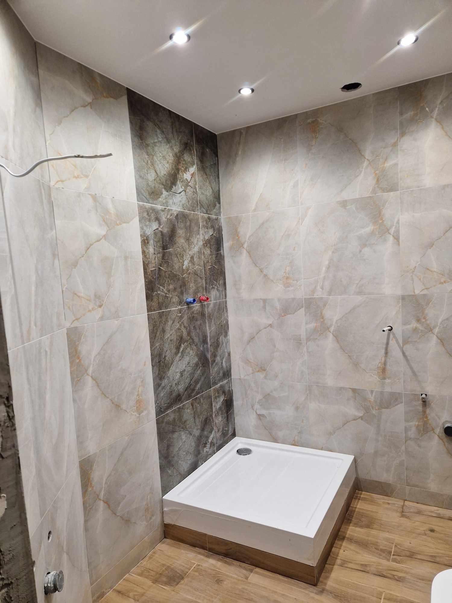 Niedokończona łazienka z marmurowymi płytkami na ścianach, widoczny biały brodzik, fragment instalacji wodnej i otwór w suficie na oświetlenie.