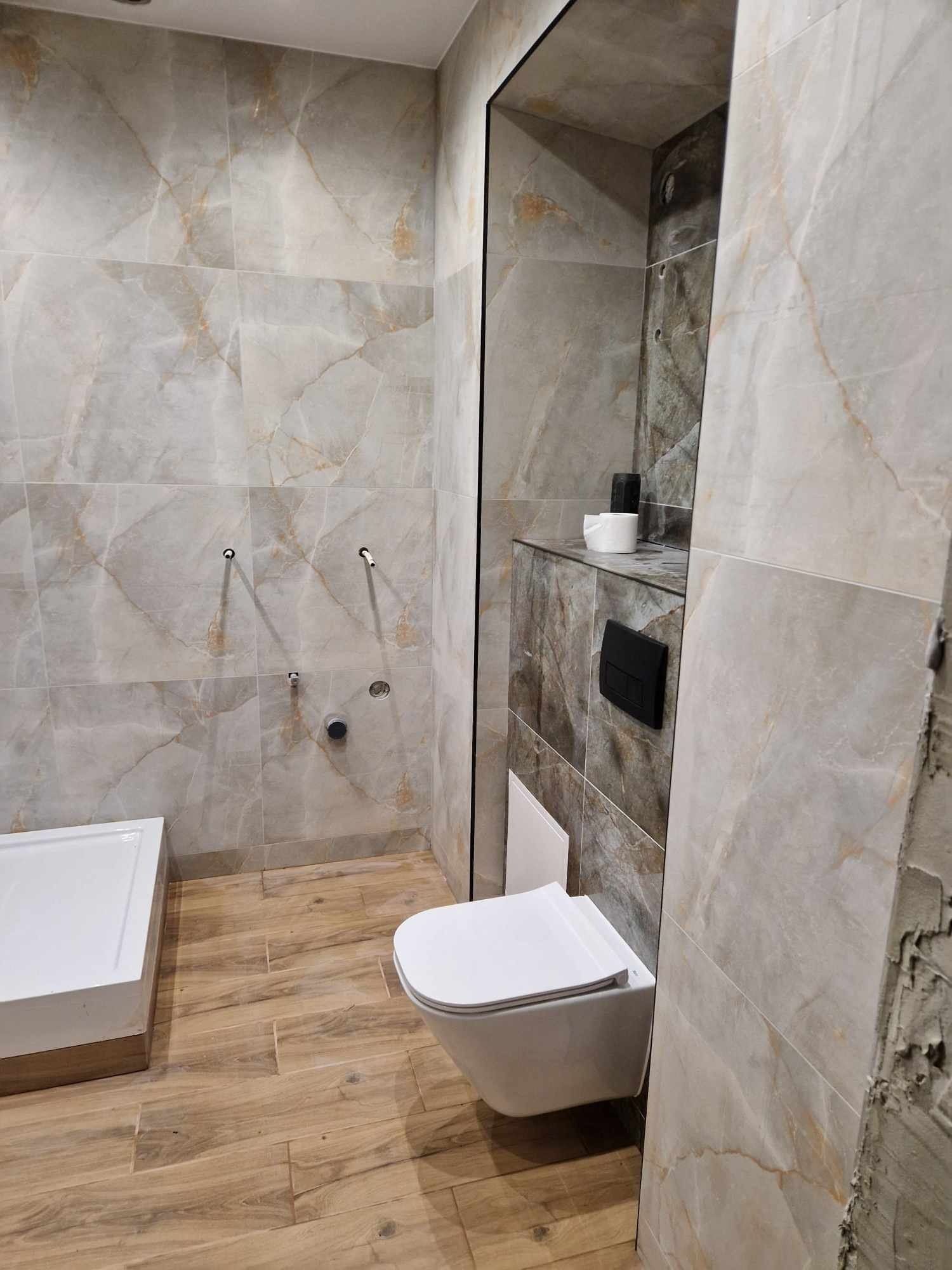 Wykończona łazienka z płytkami imitującymi marmur na ścianach, drewnopodobnymi płytkami na podłodze, białą toaletą podwieszaną i wnęką z półką. Widoczne elementy instalacji wodnej i fragment ściany...