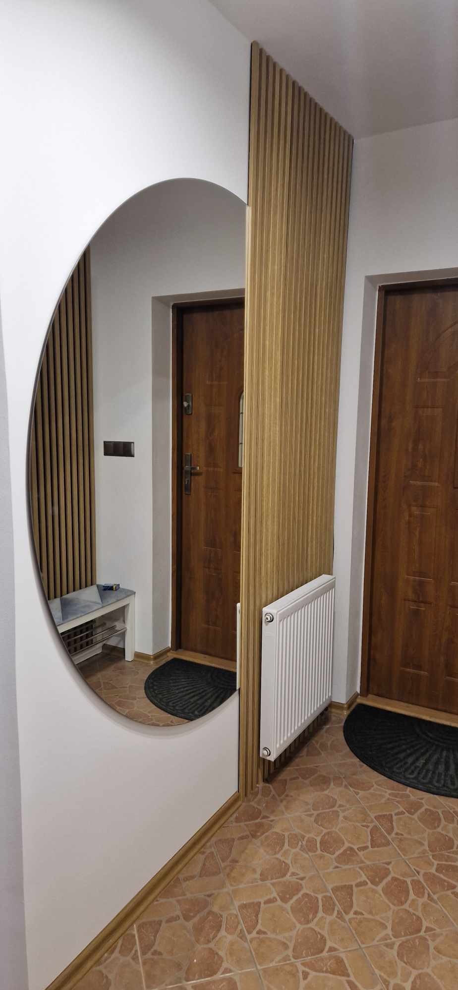 Aranżacja przedpokoju z owalnym lustrem, drewnianymi lamelami na ścianie, grzejnikiem i ceramiczną podłogą imitującą kamień.