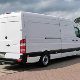 Biały samochód dostawczy Mercedes-Benz Sprinter z widocznym logo, zaparkowany na kostce brukowej na tle pochmurnego nieba i zieleni.