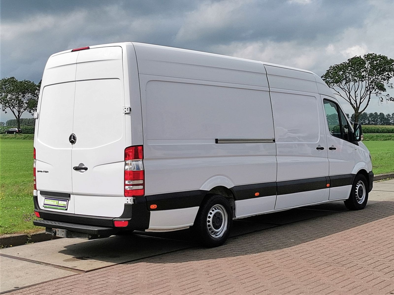 Biały samochód dostawczy Mercedes-Benz Sprinter z widocznym logo, zaparkowany na kostce brukowej na tle pochmurnego nieba i zieleni.