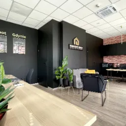 Jasne biuro agencji nieruchomości z drewnianymi biurkami, czarnymi ścianami z grafikami Gdańska, Sopotu i Gdyni, roślinami doniczkowymi i logo firmy Property Gold.