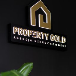 PROPERTY GOLD - Podświetlany szyld 'Property Gold Agencja Nieruchomości' z logo domu na czarnym tle, z fragmentem rośliny w dolnym rogu. Elegancki design.