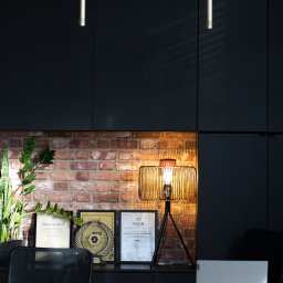 PROPERTY GOLD - Biuro z dyplomami i nagrodami na tle ceglanej ściany i czarnych szafek. Na biurku lampa i laptop. Elementy wystroju wnętrza biura nieruchomości.