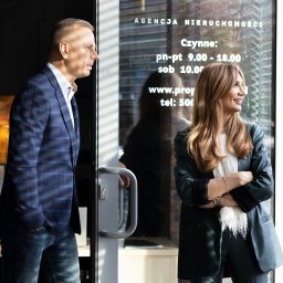 PROPERTY GOLD - Dwoje agentów nieruchomości, mężczyzna w niebieskiej marynarce i kobieta w zielonym garniturze, stoją w biurze agencji, patrząc przez okno z godzinami otwarcia.