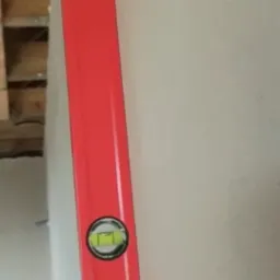 Czerwona poziomica przyłożona do świeżo położonego, jasnego tynku na ścianie w trakcie prac wykończeniowych.