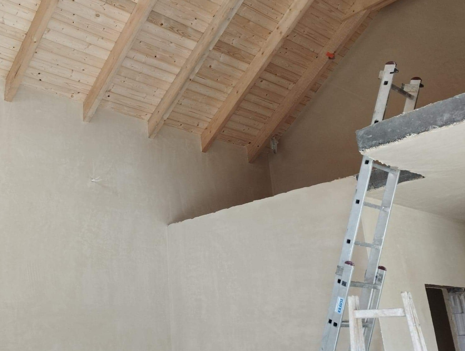 Wnętrze w trakcie prac wykończeniowych: ściany pokryte świeżym, jasnym tynkiem, drewniana konstrukcja dachu oraz aluminiowa drabina Krause.