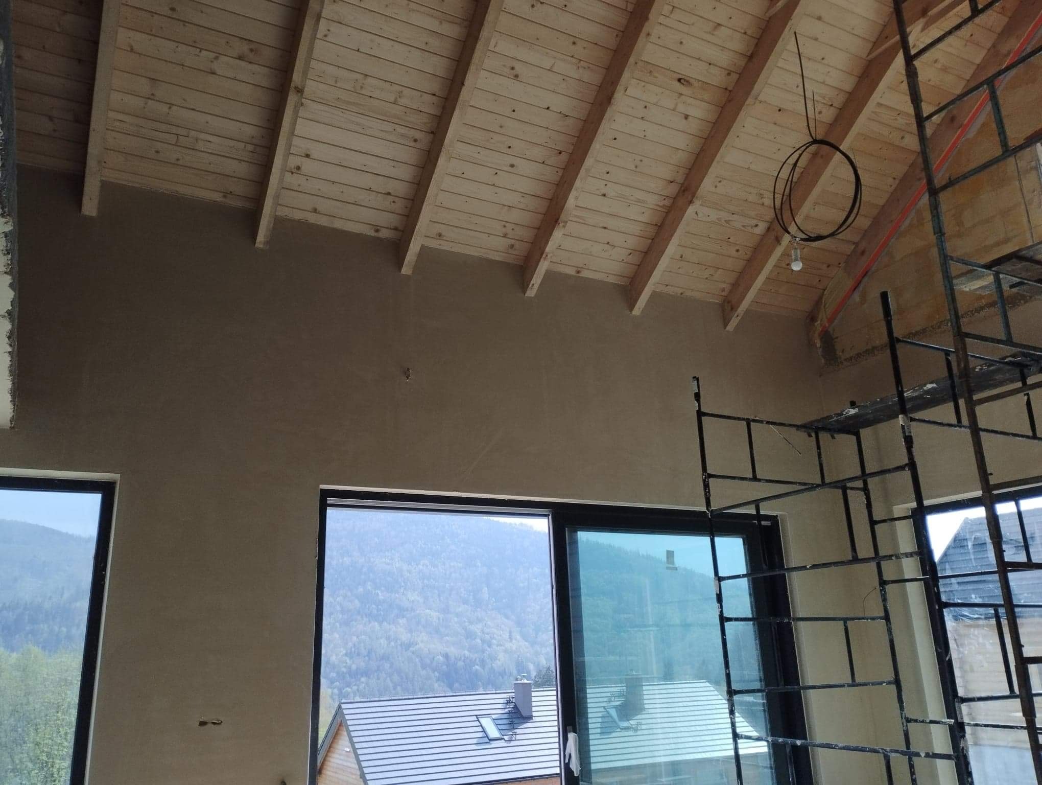 Wnętrze domu w trakcie prac wykończeniowych: ściana pokryta świeżym, jasnobrązowym tynkiem, drewniany sufit z widocznymi belkami, okna z widokiem na zalesione wzgórza, rusztowanie w rogu...