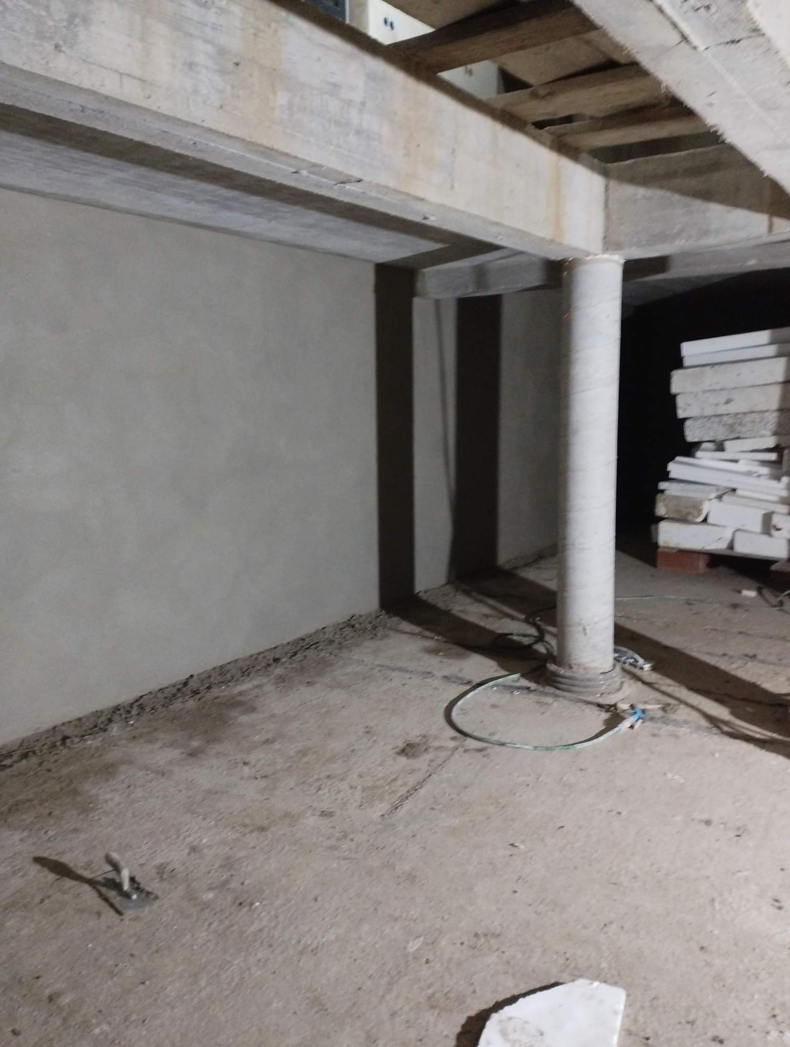 Świeżo położony tynk maszynowy na ścianie w piwnicy, widoczny filar, fragment węża i narzędzie do tynkowania na podłodze.