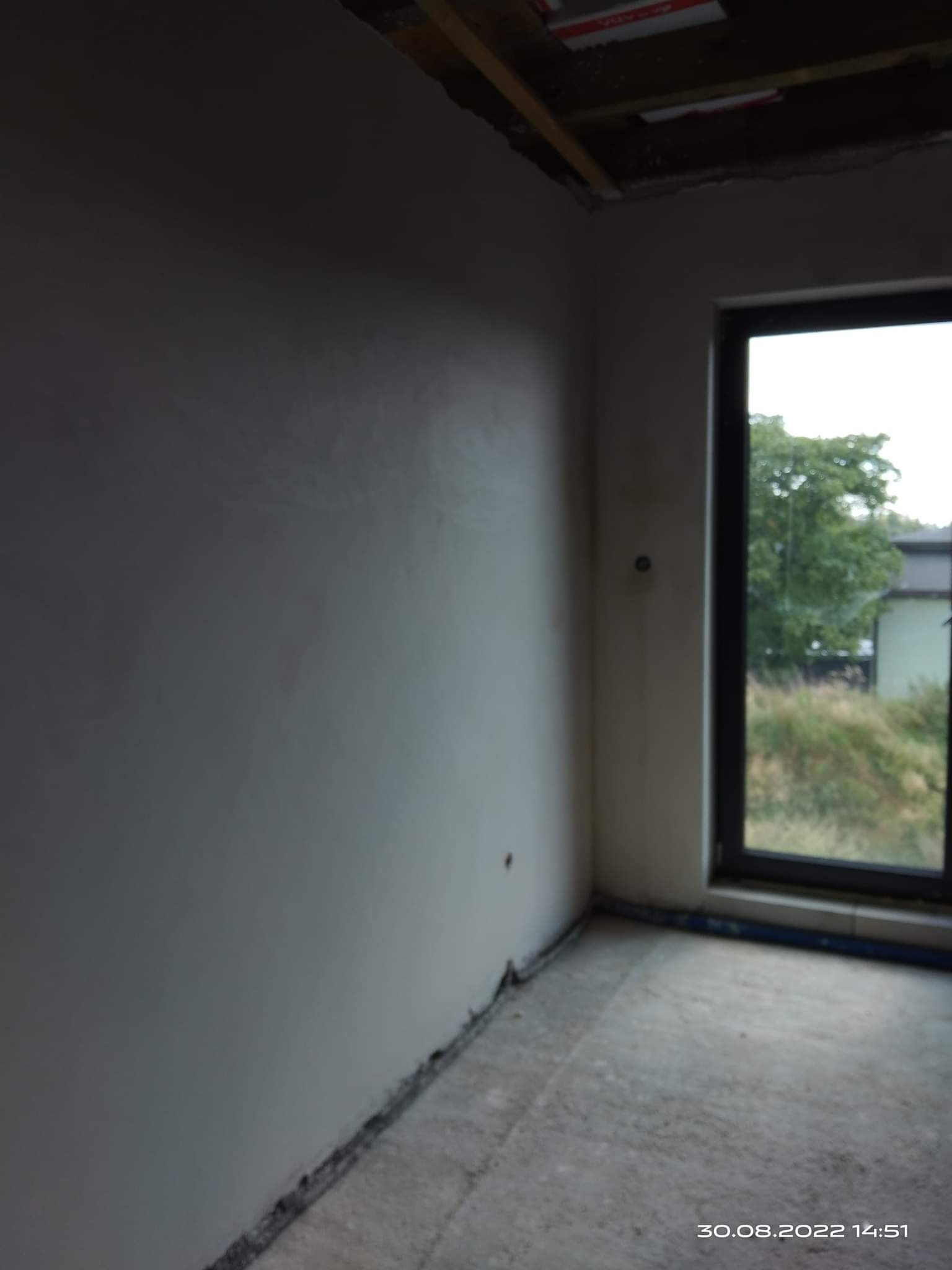 Świeżo nałożony, gładki tynk maszynowy na ścianie w surowym wnętrzu, widoczne ramy okienne i niedokończony sufit.