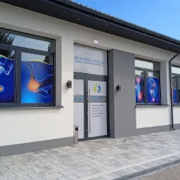 Fasada budynku centrum medycznego z napisem 'Rehabilitacja' nad wejściem, okna oklejone grafikami przedstawiającymi układ kostny i stawy, chodnik z kostki brukowej.