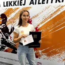 Kobieta trzymająca legitymację instruktora Polskiego Związku Lekkiej Atletyki i dyplom PZLA na tle baneru z logo PZLA.