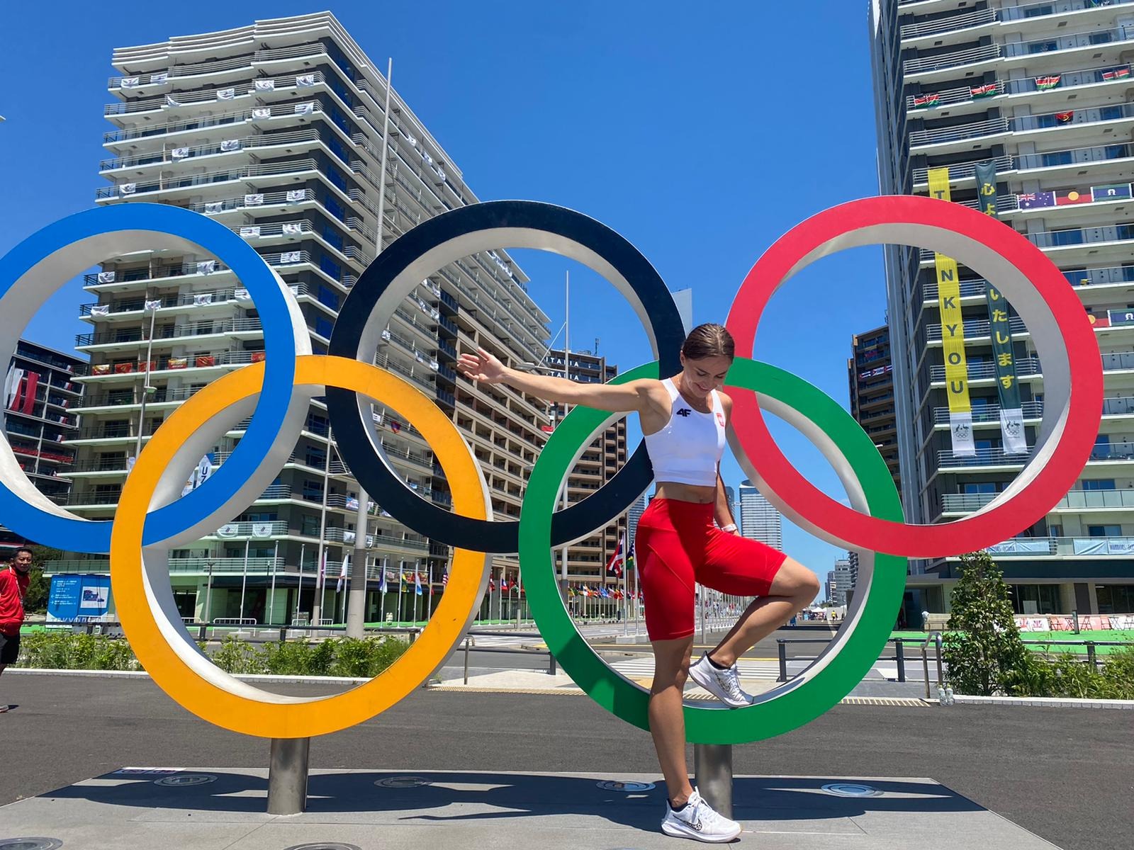 Uśmiechnięta kobieta w sportowym stroju pozuje na tle kolorowych kół olimpijskich, z budynkami i flagami w tle, w słoneczny dzień.