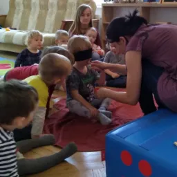 Dzieci siedzą w kręgu na podłodze, jedno z zawiązanymi oczami dotykane przez opiekunkę, w tle widoczna kanapa i dekoracje dziecięce.