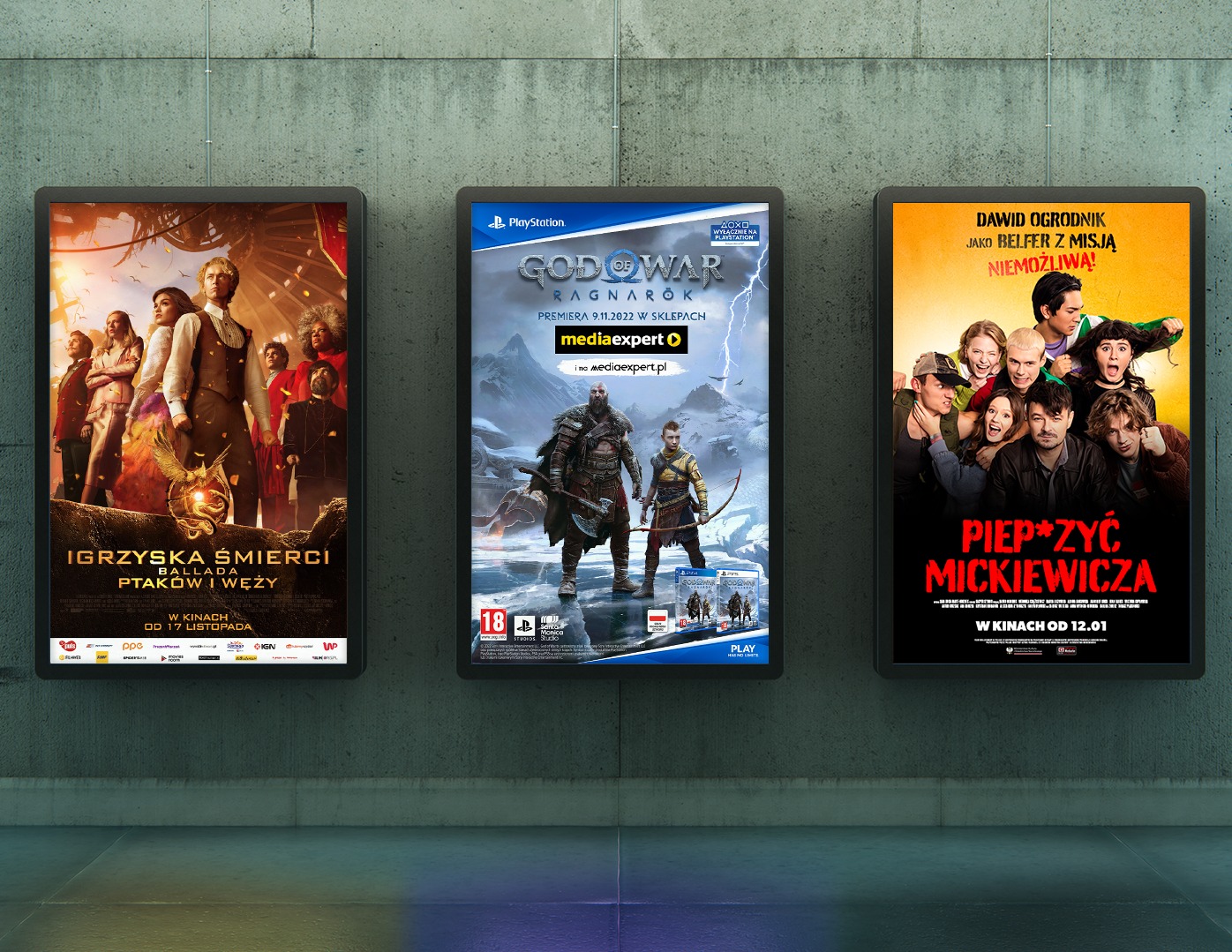 Trzy podświetlane plakaty reklamowe na ścianie: Igrzyska Śmierci, God of War Ragnarok i Piep*rzyć Mickiewicza, z datami premier kinowych i sklepowych.