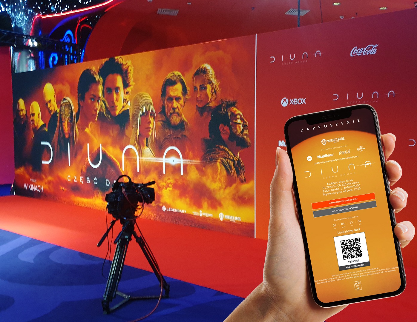 W Mińsku Mazowieckim, osoba trzyma smartfon z cyfrowym zaproszeniem na premierę filmu 'Diuna: Część Druga' na tle plakatu filmowego i kamery filmowej na statywie.