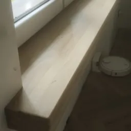 Jasny, drewniany element zamontowany pod oknem z widokiem na zieleń, obok robota sprzątającego na parkiecie ułożonym we wzór jodełki.