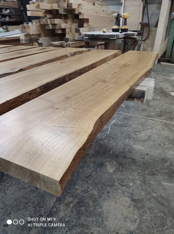 Surowe deski z naturalną krawędzią, prawdopodobnie dębowe, ułożone w warsztacie, z widocznym stosem desek w tle i wkrętarkami na stole.