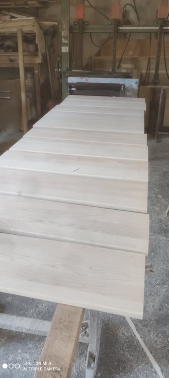 Surowe, jasne deski ułożone na drewnianym stojaku w warsztacie, przygotowane do dalszej obróbki, z widoczną maszyną stolarską w tle.