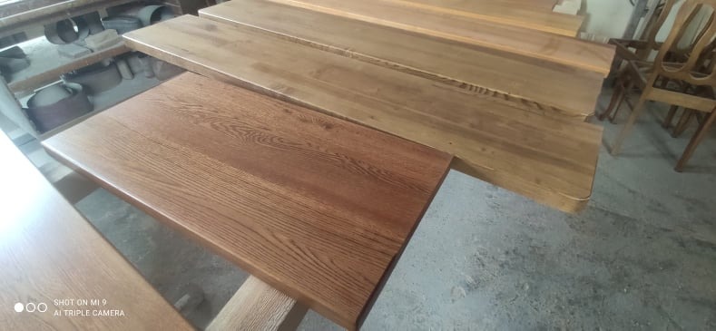 Przygotowane do montażu drewniane elementy, prawdopodobnie blaty, w warsztacie, z widoczną strukturą drewna i różnicami w odcieniach bejcy.