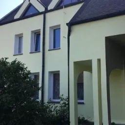 Dwupiętrowy budynek z jasną elewacją, widoczne okna z białymi ramami, ciemny dach i fragment zadaszonego wejścia z łukami.