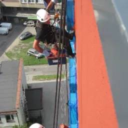 Dwóch alpinistów przemysłowych, zabezpieczonych linami, maluje pomarańczową elewację budynku, z widocznymi zabezpieczeniami okien folią w kolorze niebieskim, z perspektywy z dołu w słoneczny dzień.