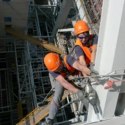 Dwóch monterów w kaskach i kamizelkach, zabezpieczonych linami, wierci w metalowej konstrukcji na wysokości, w tle skomplikowana konstrukcja stalowa.