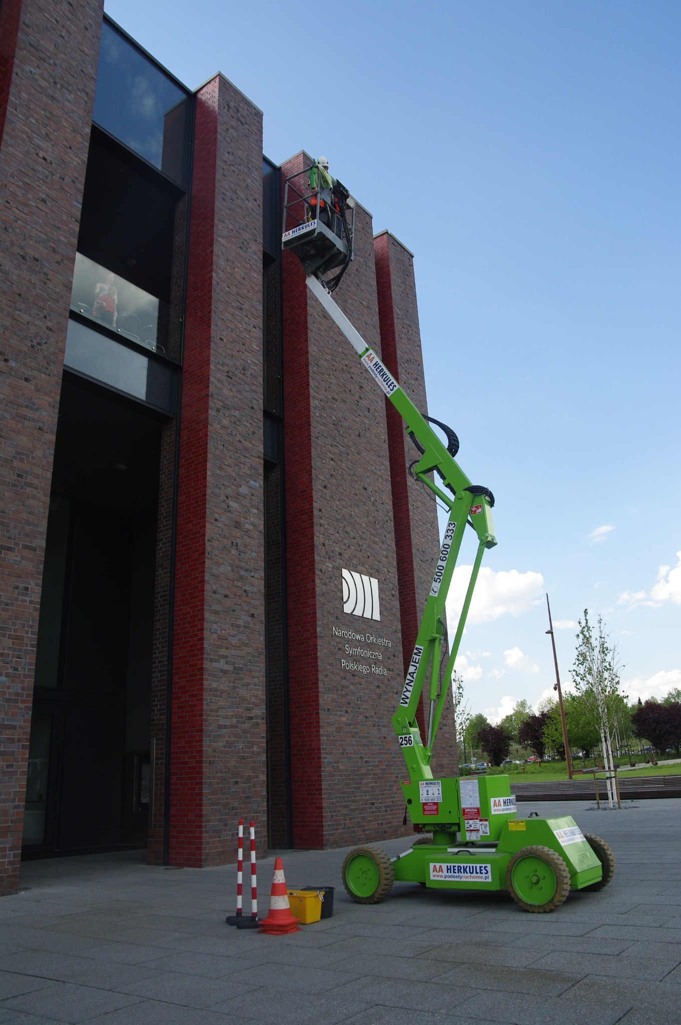 Pracownik w koszu podnośnika teleskopowego na tle budynku Narodowej Orkiestry Symfonicznej Polskiego Radia, widoczne zabezpieczenia i oznakowanie terenu.
