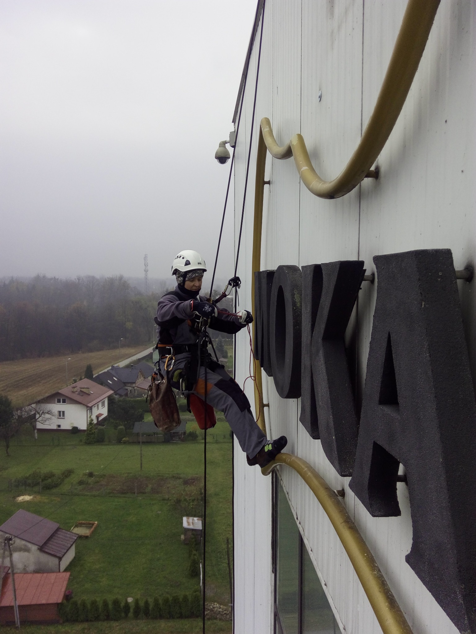 Pracownik na linach zabezpieczających, w kasku i uprzęży, czyści lub naprawia duży, czarny napis 'ROKA' na białej elewacji budynku. W tle widoczny krajobraz z domami i drzewami w mglisty dzień.