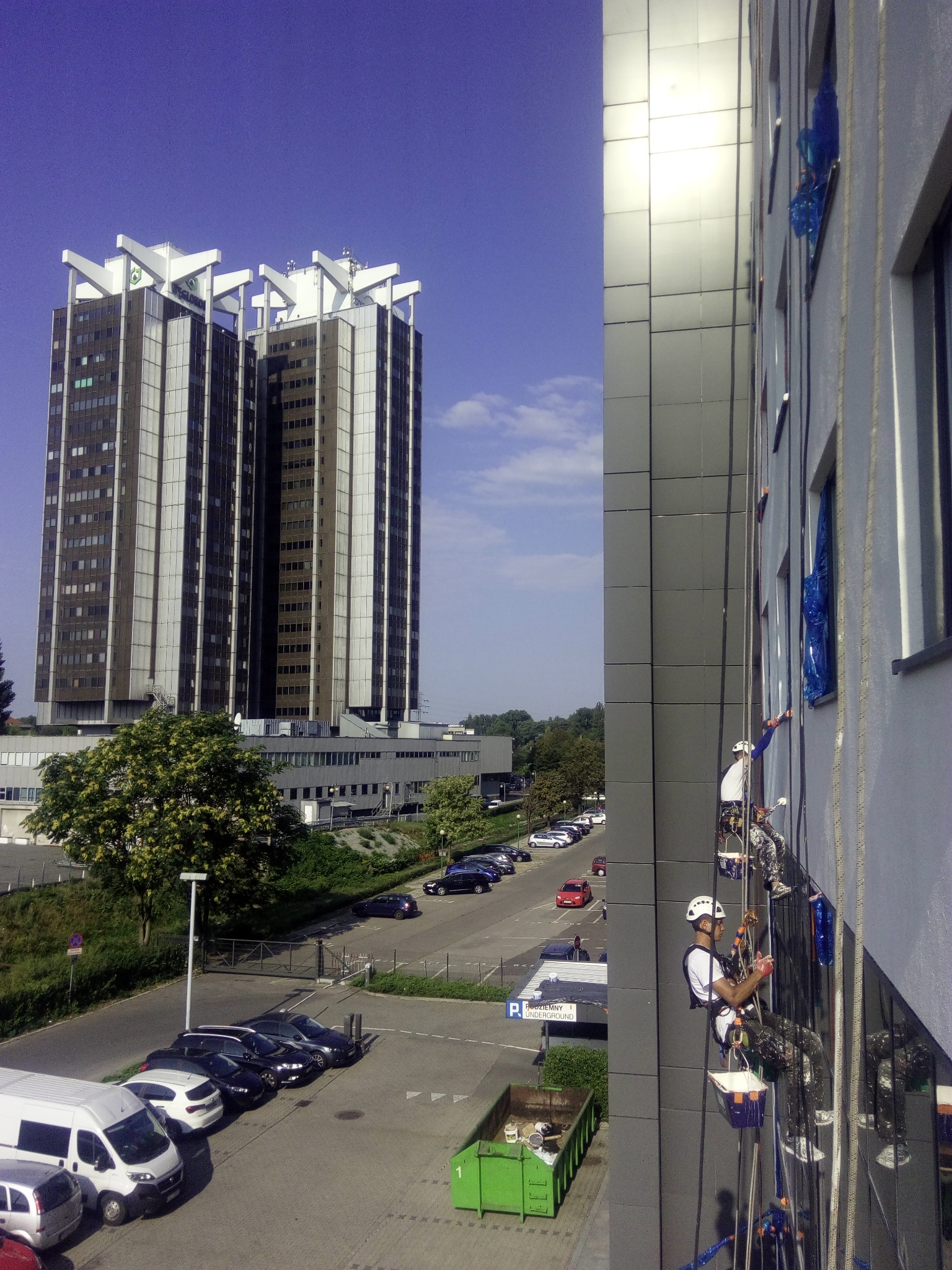Pracownicy na linach zabezpieczających, podczas prac na wysokościach przy nowoczesnym biurowcu z szarymi panelami, widoczne zabezpieczenia okien folią, w tle drugi wysoki budynek, parking i zieleń.