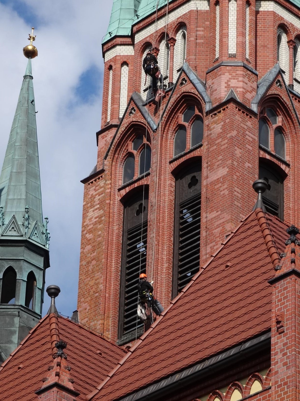 Dwóch alpinistów przemysłowych na linach, pracujących na ceglanej wieży kościelnej z zielonym dachem, widoczny fragment dachu z czerwonej dachówki.