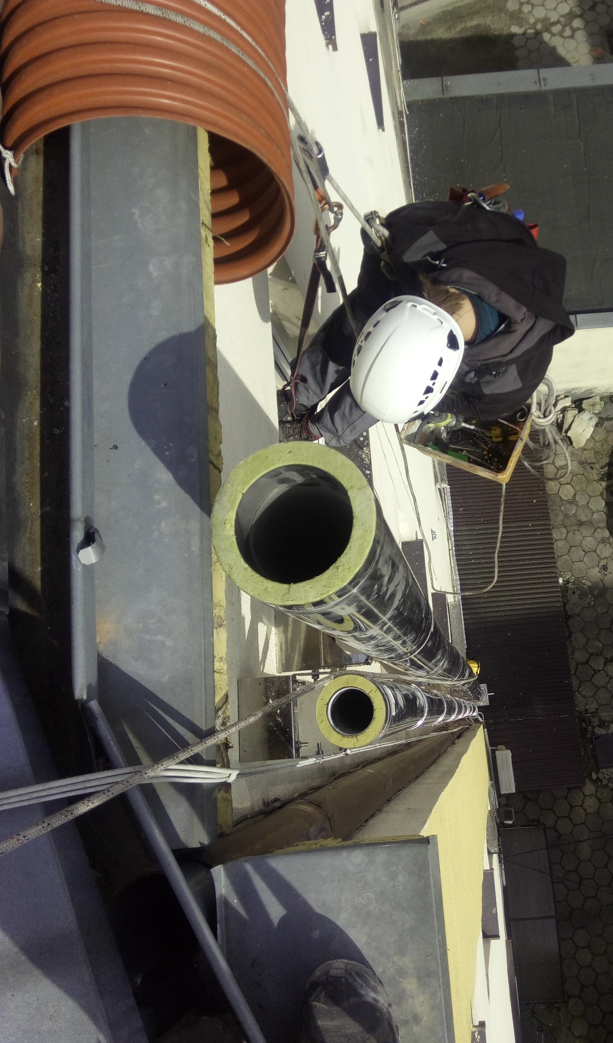 Pracownik w kasku, zabezpieczony linami, montuje izolowane rury na zewnątrz budynku, widok z góry.