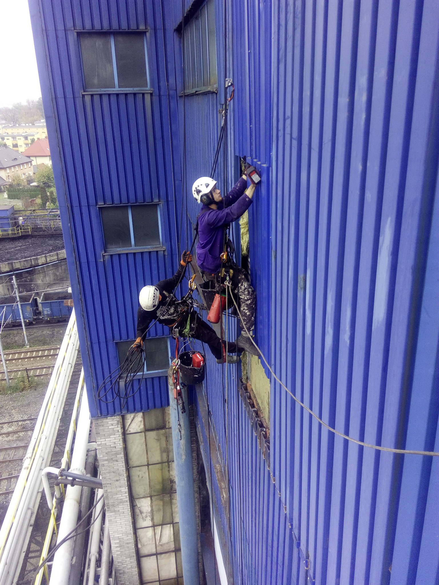 Dwóch pracowników na linach zabezpieczających, jeden naprawia uszkodzony fragment niebieskiej, blaszanej ściany budynku przemysłowego, drugi trzyma przewody, w tle widoczne zabudowania i tory...