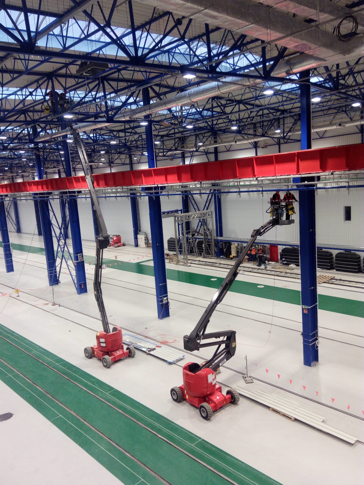 Wnętrze hali przemysłowej z dwoma podnośnikami koszowymi; pracownicy w kaskach na wysokości, montaż instalacji pod sufitem z widoczną konstrukcją stalową i świetlikami.