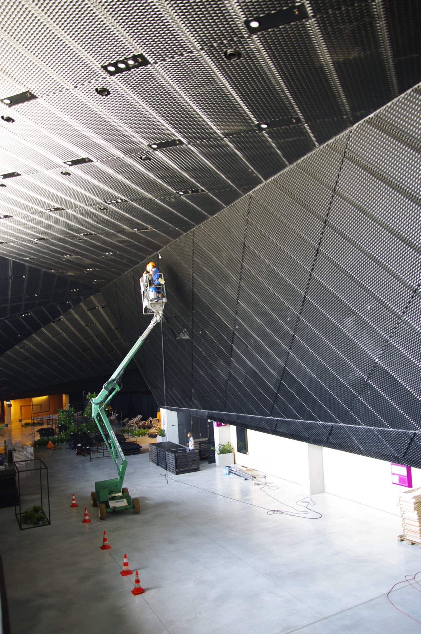 Pracownik na podnośniku koszowym przy metalowej konstrukcji sufitu w dużej hali, widoczne stożki ostrzegawcze na podłodze.