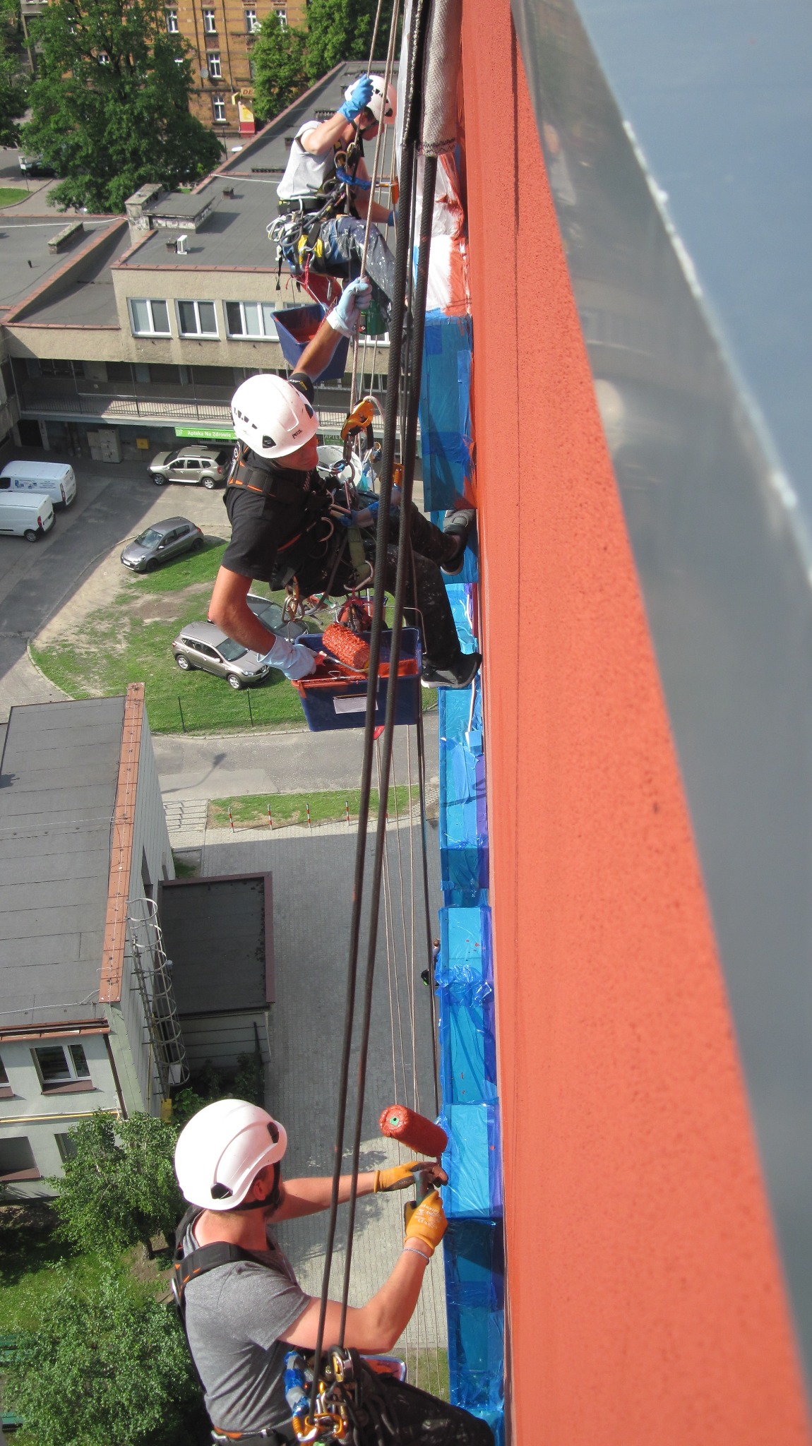 Dwóch alpinistów przemysłowych, zabezpieczonych linami, maluje pomarańczową elewację budynku, z widocznymi zabezpieczeniami okien folią w kolorze niebieskim, z perspektywy z dołu w słoneczny dzień.