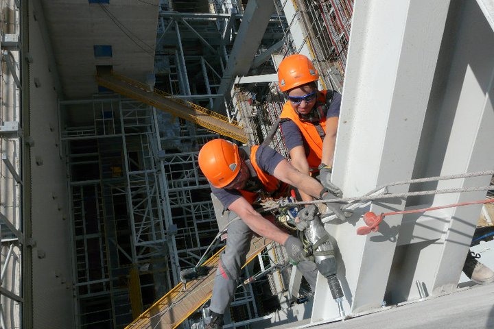 Dwóch monterów w kaskach i kamizelkach, zabezpieczonych linami, wierci w metalowej konstrukcji na wysokości, w tle skomplikowana konstrukcja stalowa.