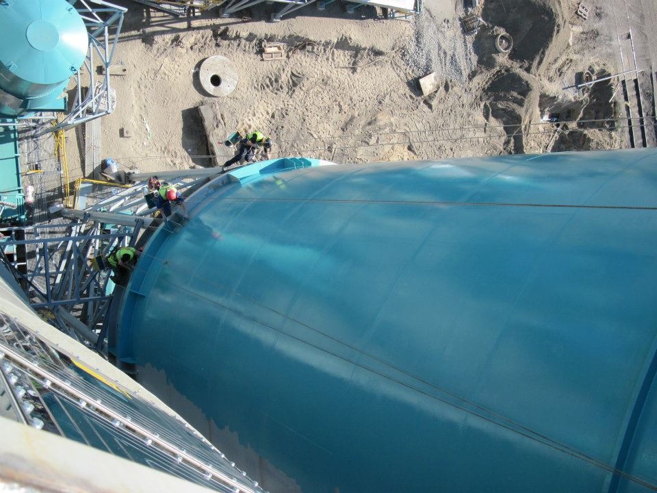 Prace wysokościowe przy malowaniu dużego, turkusowego zbiornika przemysłowego z widocznymi pracownikami w kaskach i uprzężach, ujęcie z góry.