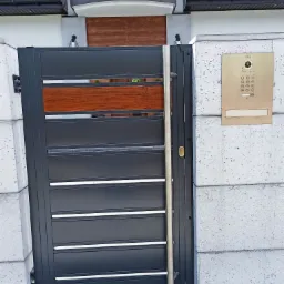 Nowoczesna brama wjazdowa z poziomymi panelami w kolorze grafitowym, z elementami dekoracyjnymi z drewna, zamontowana przy betonowym słupku i wyposażona w wideodomofon.