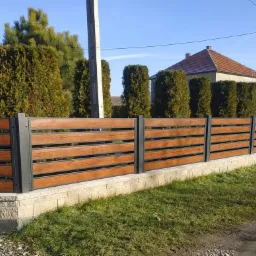 Nowoczesne ogrodzenie z poziomych desek o barwie drewna, osadzone na szarych, metalowych słupkach i niskim, kamiennym murku, na tle zielonego trawnika i zabudowań.