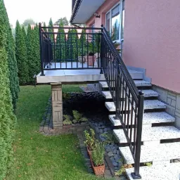 Metalowa balustrada schodowa w kolorze czarnym, prowadząca do wejścia domu z różową elewacją, z widokiem na ogród z tujami i kamienną aranżacją pod schodami.