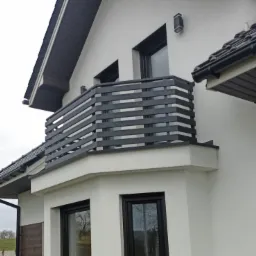 Nowoczesny balkon z szarymi, poziomymi panelami na białym domu z czarnymi ramami okiennymi i ciemnoszarą dachówką.